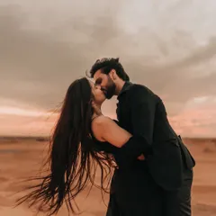 A passionate kiss in the Dubai desert as the wind catches her hair. This image is full of movement and emotion, set against the warm, dramatic colors of a desert sunset.