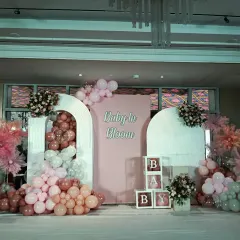 A large and elegant "Baby in Bloom" stage decoration. This setup uses modern arch backdrops, a soft pink color scheme, and beautiful floral arrangements mixed with balloons for a sophisticated look.