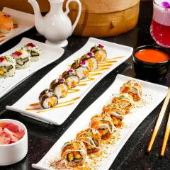 A vibrant spread of assorted sushi rolls, elegantly plated on long white dishes. The frame includes a bamboo steamer and other props to create a complete Asian dining scene, perfect for a restaurant menu.
