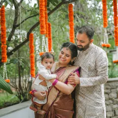 A lovely family portrait taken outdoors during a naming ceremony. The parents embrace their child under hanging marigold flower arrangements, creating a warm and traditional photograph.