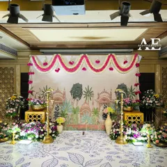 An elegant and artistic stage for an engagement. The backdrop is a printed canvas with a palace motif, framed by fresh flower garlands and traditional brass lamps.