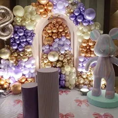 An elaborate teddy bear themed setup featuring a stunning balloon arch in metallic purple, gold, and cream. The addition of a large, stylized bear figure and matching pedestals elevates the theme into a luxurious and modern celebration.