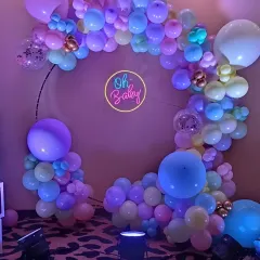 Pastel balloon rings are a beautiful and soft option for a baby shower. This setup uses a mix of purple, blue, yellow, and pink balloons with an 'Oh Baby' neon sign at the center.