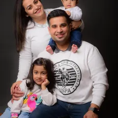 A beautiful family portrait showcasing coordinated outfits against a classic studio backdrop. The baby sitting on dad's shoulders adds a lovely, playful touch.