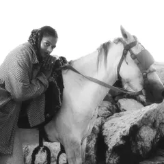 A powerful black and white fashion shot of a model leaning against a white horse amidst a rocky landscape. The high-contrast lighting and strong composition create a bold, editorial feel.