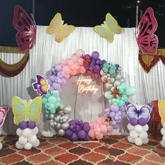 An outdoor butterfly-themed setup with a pastel balloon ring, neon sign, and large butterfly cutouts on balloon pillars.