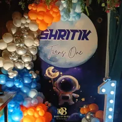 A space-themed first birthday party for a little astronaut. The decor features a personalized backdrop with a moon and astronaut graphic, framed by balloons in orange, blue, and silver, and a light-up number '1'.