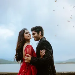A candid moment of connection as petals fall around the couple during their lakeside pre-wedding session. The woman's vibrant red gown stands out beautifully against the soft sky.