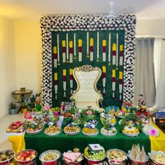 This is a housewarming ceremony setup featuring a backdrop made of banana leaves and flowers. The table is filled with my decorative platters holding sweets, fruits, and other traditional items.