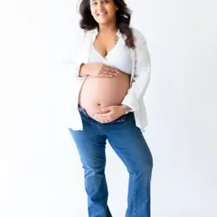 A joyful, front-facing portrait of a mother-to-be, comfortably dressed in jeans. The bright, even lighting in the studio creates a fresh and happy feel for this maternity photo.