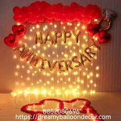 A beautiful anniversary backdrop featuring a wall of fairy lights. The 'Happy Anniversary' foil balloons are surrounded by red balloons and a heart made of rose petals and candles on the floor.