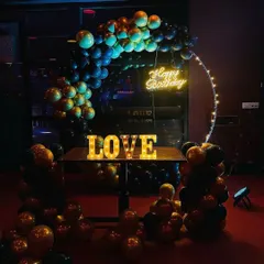 We can adapt our decorations to suit the mood of your venue. In this dimly lit space, a black and gold balloon arch with a 'Happy Birthday' neon sign and 'LOVE' marquee letters creates an intimate and celebratory atmosphere.