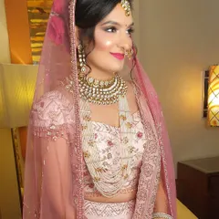 A profile shot of a bride in a delicate pastel pink lehenga. Her makeup is soft and romantic, with a pink lip and glowing skin, complemented by a heavy kundan necklace and a simple maang tikka.