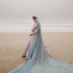 A bride stands on a serene beach at dusk, her magnificent light blue veil trailing behind her, creating a breathtaking and ethereal bridal portrait against the ocean.