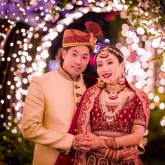 A classic portrait of a couple in stunning traditional wedding attire, framed by a beautiful arch of fairy lights. The warm glow highlights the intricate details of their outfits.