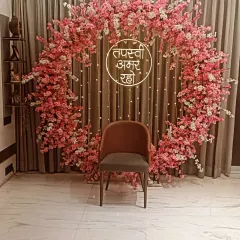 A circular arch made entirely of pink cherry blossoms for a Jain ceremony. This setup provides a delicate and beautiful frame for the main seat, complemented by a neon "Tapasvi Amar Raho" sign.