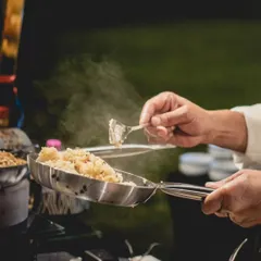 A chef serves up a freshly tossed dish from a steaming pan at an outdoor event. Our live stations are designed to handle any setting, ensuring your guests enjoy hot, gourmet food made to order.
