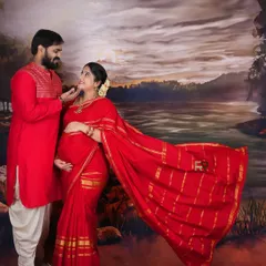 This portrait captures a couple in matching red traditional outfits against a painted landscape backdrop. The flowing saree adds a touch of drama and elegance to the scene.