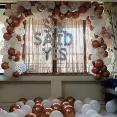 She said yes. This engagement announcement decor features a classic balloon arch in white and metallic bronze, with silver foil letters and a fringe curtain backdrop to celebrate the special moment.
