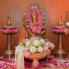 A close up of a Ganpati idol adorned with a fresh pink flower garland. The setup includes traditional brass lamps (samai) and a bowl of lotuses, creating a simple yet beautiful space for worship.