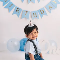 A simple birthday setup with a "Happy Birthday" banner and a party hat. The focus remains entirely on the baby's adorable, expressive face.