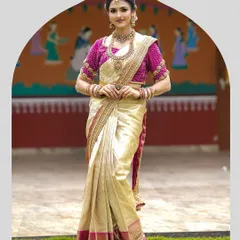 A full-length view of a classic South Indian bridal look. The bride wears a cream and gold silk saree with a richly embroidered magenta blouse, standing against a backdrop that evokes traditional charm.