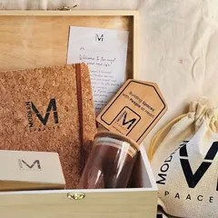 An open view of a 'Modern Spaaces' welcome kit. It includes a branded cork notebook, a glass jar, a welcome note, and other essentials packed in a custom wooden box.