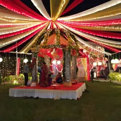 A vibrant wedding mandap under a canopy of red, white, and yellow drapes, with decorated pillars and ample lighting for a nighttime ceremony.