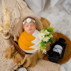 A sleeping newborn is nestled in a basket with a floral headband, set against a soft, bohemian-themed backdrop with dried flowers and macrame.