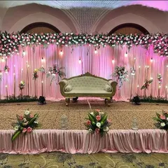Here is a reception stage setup with a soft, romantic feel. I used a backdrop of pink drapes with hanging bulb lights, topped with a dense arrangement of pink and white flowers to create a warm and inviting space.