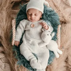 A sleeping newborn in a cozy, textured white romper and matching hat, holding a small knitted toy. This boho-inspired shot uses natural textures like pampas grass for a warm, earthy feel.