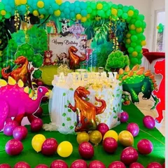A prehistoric adventure with this fun dinosaur-themed birthday party. The setup includes a lush green backdrop, various dinosaur cutouts and foil balloons, and a balloon-wrapped cake table.