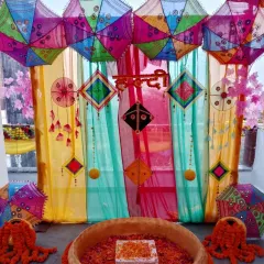 A vibrant Haldi ceremony space featuring a multi-colored backdrop with hanging kites and umbrellas. The 'urli' is filled with flower petals, ready for the rituals.