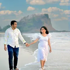 A candid moment of a couple walking hand-in-hand along the water's edge. The bright, sunny day and the distant coastal mountain create a beautiful, natural setting for this pre-wedding photograph.