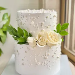 A vision in white, this two-tiered cake is the definition of timeless elegance. The clean buttercream finish is adorned with scattered edible pearls and fresh white roses, making it a perfect centerpiece for a sophisticated roka or wedding celebration.