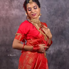 A studio session featuring a model in a vibrant red traditional saree. The mottled grey backdrop ensures the rich colors and gold jewelry stand out.