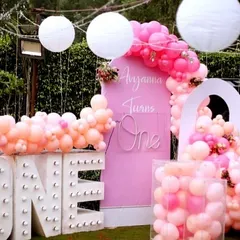 A beautiful pink-themed first birthday setup outdoors, with a large light-up 'ONE', a cascade of pink and peach balloons, and paper lanterns.
