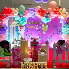 A fairy and butterfly themed second birthday party. This colorful setup includes a purple backdrop, a rainbow balloon arch, and cutouts of whimsical fairies.