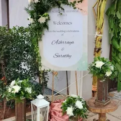 A rustic and elegant welcome sign for Ashrraya and Sarathy's wedding, framed with fresh white flowers and greenery, and accented with lanterns.