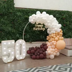A sophisticated backdrop designed for a 50th birthday, featuring a balloon ring with a gradient of white, caramel, and chocolate tones. The setup is completed with large, illuminated "50" marquee numbers and a "Happy Birthday" neon sign against a lush green wall.