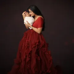 A full-length portrait of a new mother in a flowing red gown, holding her swaddled newborn close. The image is filled with emotion and has the feel of a classic painting.