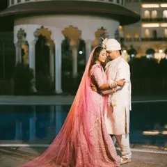 A regal portrait of the bride and groom by the poolside at night. The bride's flowing pink lehenga and the grand architecture create a stunning, cinematic image.
