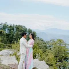 A couple shares an intimate gaze on a rocky ledge, with the soft blue sky and distant snow-capped peaks creating a serene and romantic atmosphere for their pre-wedding portrait.