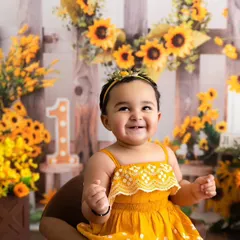 A baby girl in a yellow dress gives a huge, happy smile during her sunflower-themed first birthday photoshoot.
