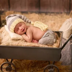 A smiling newborn wearing a striped tassel hat sleeps in a small metal wagon filled with hay, creating a charming and rustic farm-themed scene.