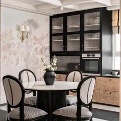 The informal dining area in Riviera Maison, where we paired a fluted-base marble-top table with classic striped dining chairs. The black and oak cabinetry and a soft landscape mural create a sophisticated, European-inspired look.