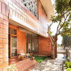 This view of the Reshma Residence highlights its modern composition, where clean lines and simple forms meet the texture of exposed brick. The design creates a small, inviting porch that connects the home directly to the garden path.