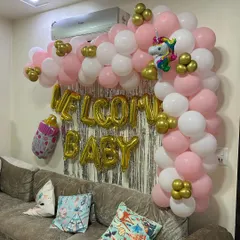 A classic "Welcome Baby" decoration for a living room. This setup features a pink, white, and gold balloon arch over the sofa, a silver foil curtain backdrop, and large gold foil letters.