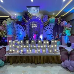 A magical butterfly-themed birthday stage in a banquet hall. The decor features large illuminated butterfly wings, a custom name sign, and a beautiful balloon arch.