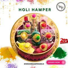 A beautifully arranged Holi hamper in a round basket, surrounded by colorful gulal. This gift set includes bottles of syrup, jars of sweets and nuts, and other festive treats, all elegantly tied with twine.